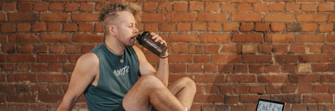 Athletic man sitting on exercise mat with water bottle, taking a break from workout, in a rustic indoor setting.